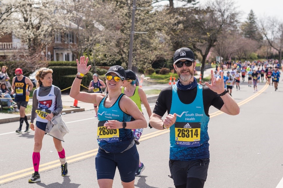 Two runners wave while making their way through the Boston Marathon route