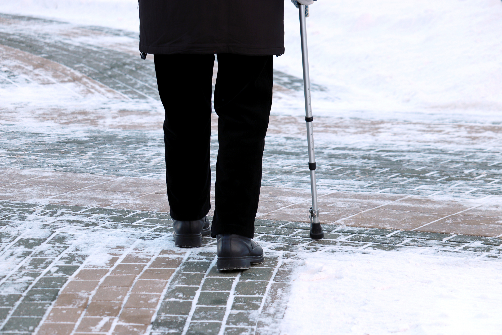 Photo of an older person using a cane walking on a brick pathway lightly covered in snow and ice