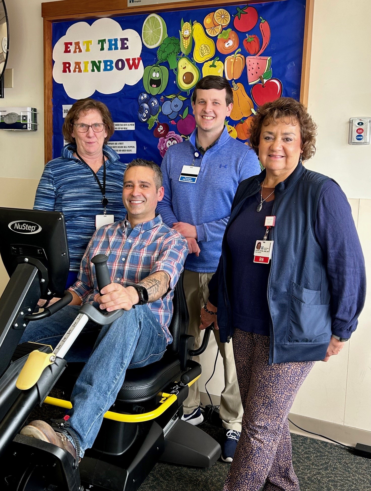 A male patient sitting on an exercise bike surrounded by three cardiac rehab team members
