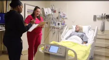 Two female nurses stand by a female patient's bedside and go over her care plan 