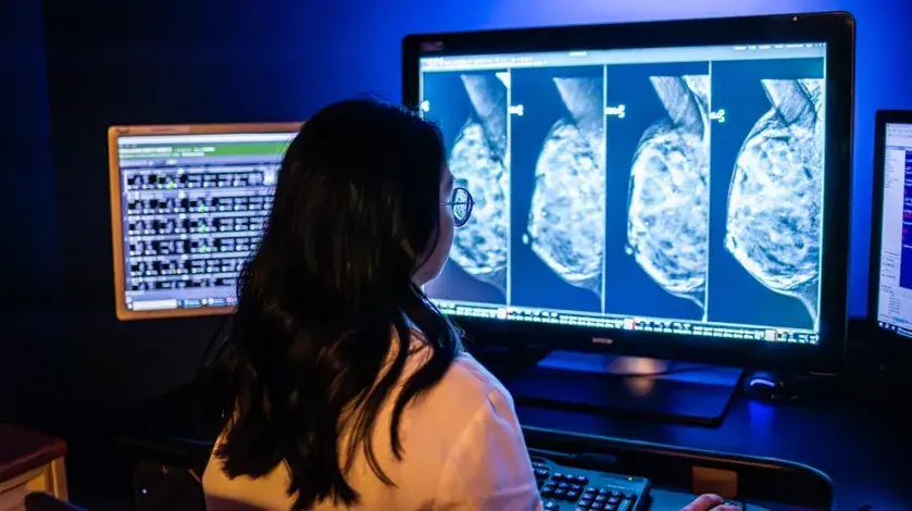 A clinician sits in a dark room looking at a computer screen that features four images of a diagnostic breast scan