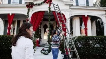 A man uses a ladder to hang Christmas lights on a house