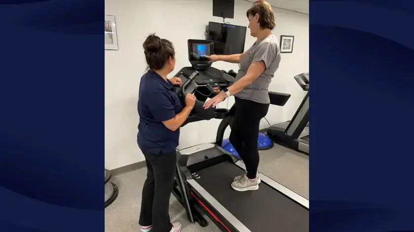 An exercise therapist points out functions on a treadmill while a class attendee stands on the treadmill listening.