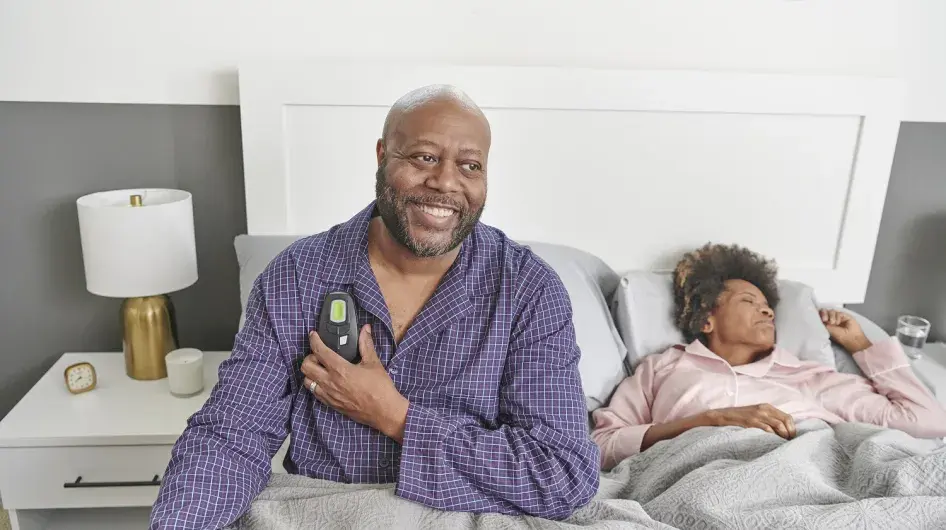 A man smiles as he sits up in bed holding his Inspire Therapy remote. His wife is asleep next to him.