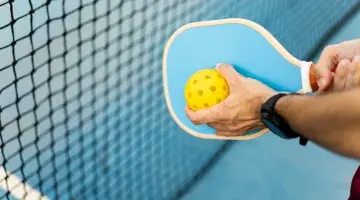 A view of a person holding a yellow pickleball in front of a blue pickleball paddle.
