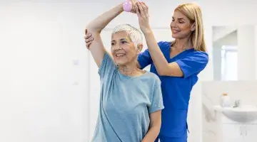Woman in scrubs helps a patient lift a weight over her head at home-based PT - 2024