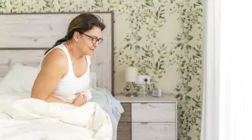 A woman sits on the edge of her bed holding her stomach with an uncomfortable expression on her face.