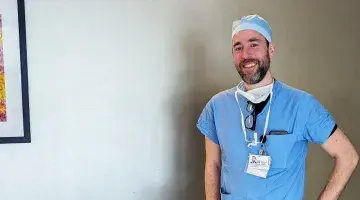 John Photos, CRNA poses for a photo wearing blue scrubs and a surgical cap