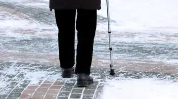 Photo of an older person using a cane walking on a brick pathway lightly covered in snow and ice