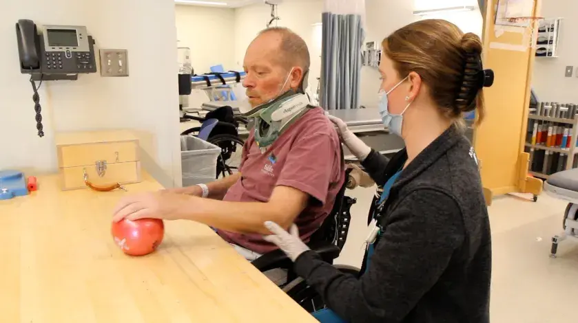 Ed sitting at a table with a physical therapist, moving a weighted ball back and forth on the table with his hand, wrist, and forearm