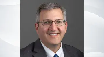 Professional headshot of Sam Skura, COO of South Shore Health, on a gray wave patterned background