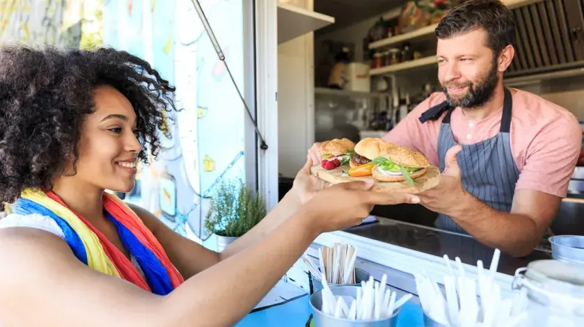 A woman smiles as a man hands her a plate of food from the window of a food truck