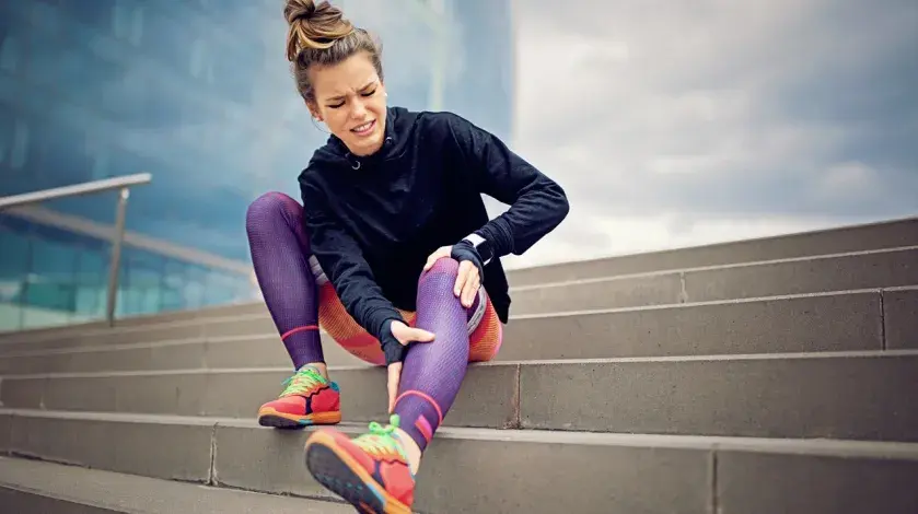 A woman wearing running clothes sits on steps holding her calf with a pained expression.