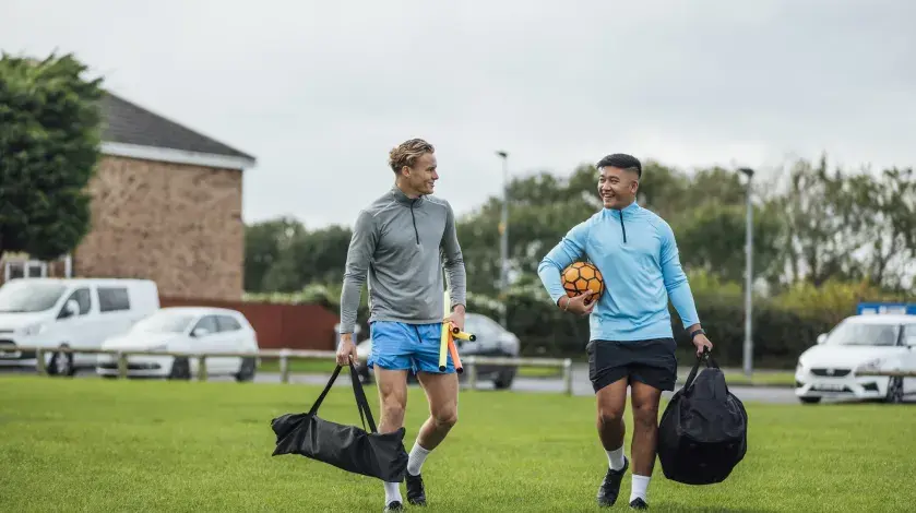 Two men smile as they talk to each other walking onto a soccer field, each carrying a bag, with balls, corner flags, etc.